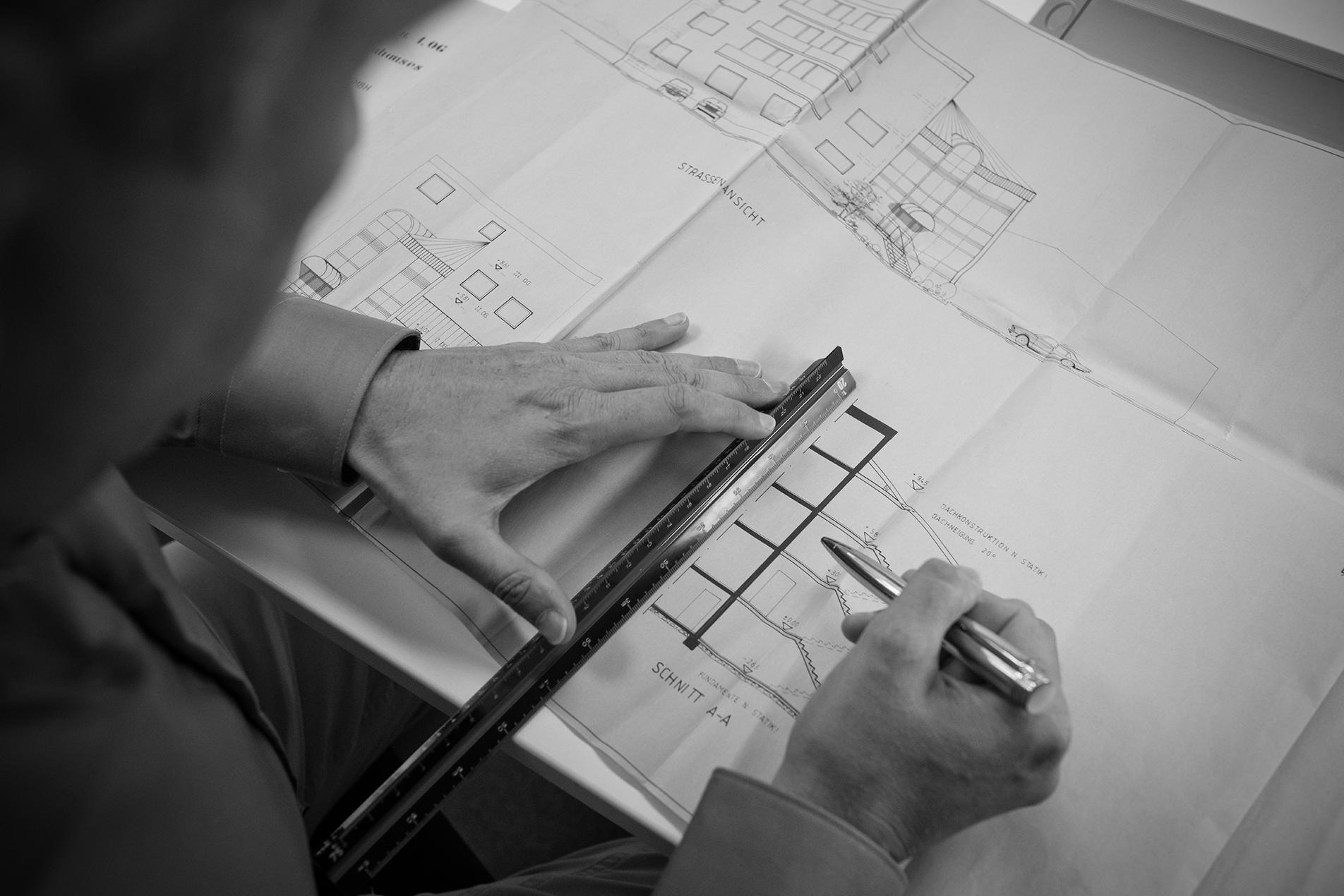 Detailaufnahme über die Schulter eines Mitarbeitenden mit Fokus auf dessen Hände, welche mit Lineal und Stift eine Achse auf einem Bauplan kontrollieren