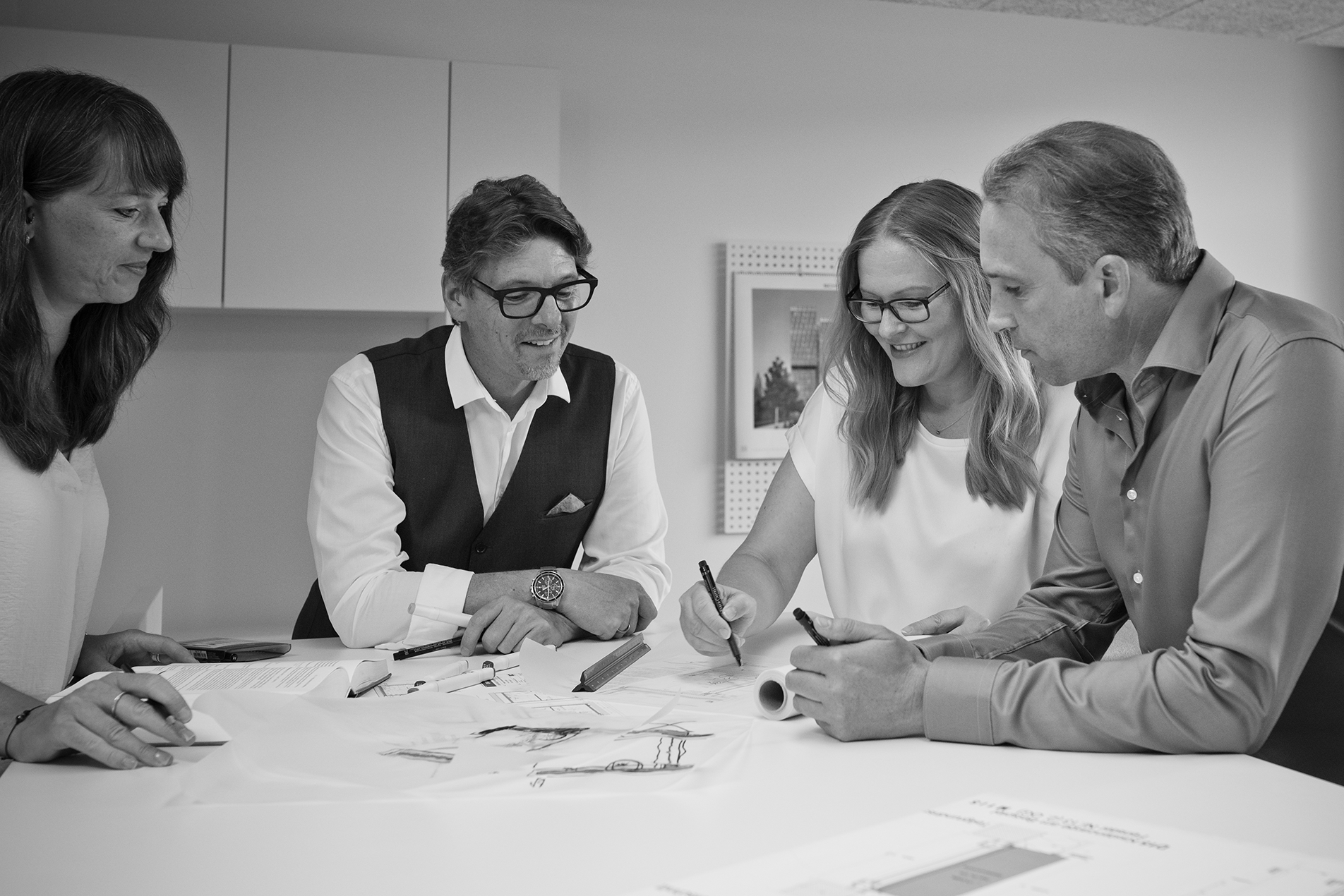 Carsten Herges und Guido Haid im Gespräch mit Mitarbeiterinnen stehend an einem Tisch. Auf dem Tisch sind diverse Skizzen, Bücher, Dokumente sowie verschiedenes Zeichen- und Schreibmaterial verteilt