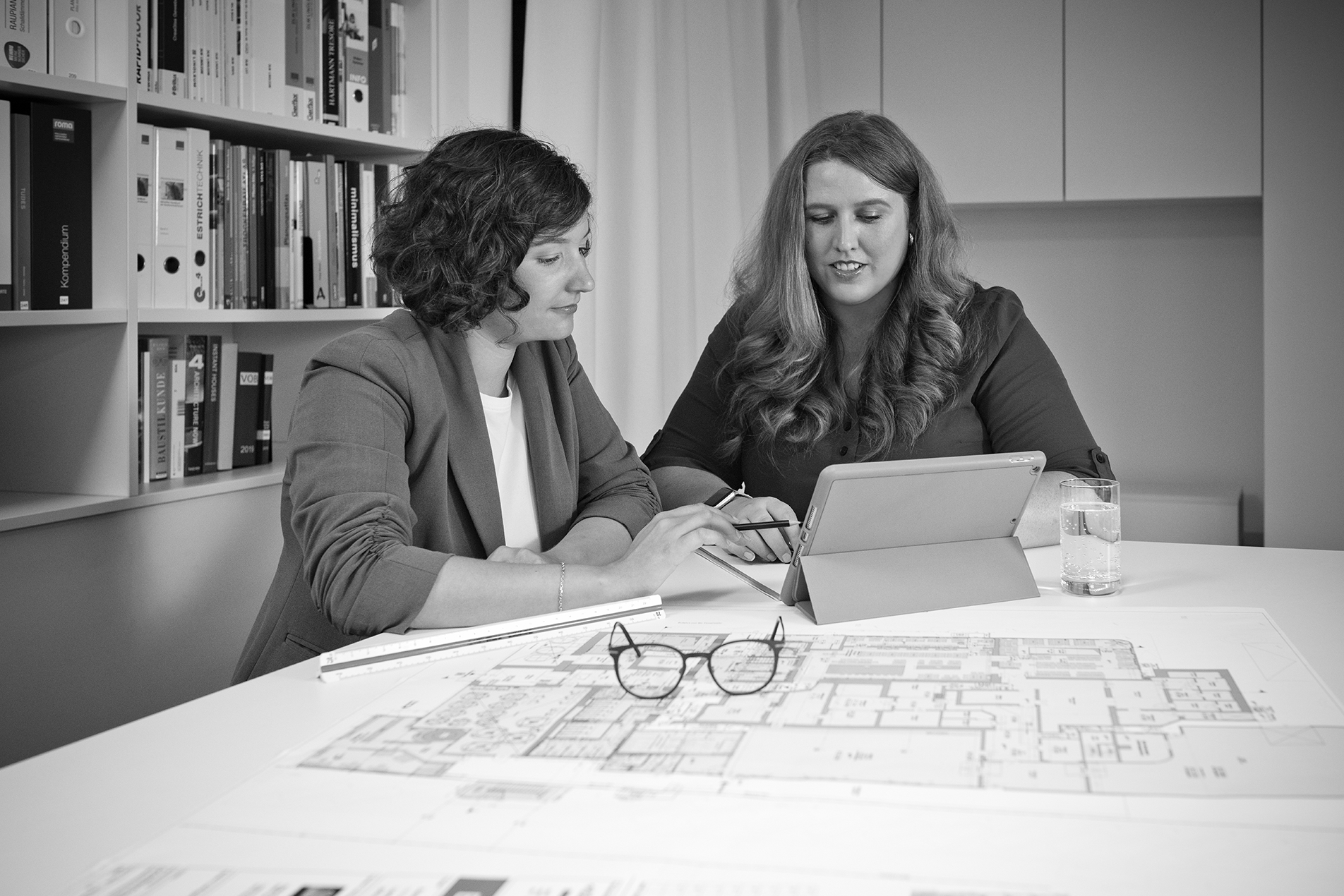 Zwei Mitarbeitende im Gespräch sitzend an einem Tisch vor einem Tablet, im Vordergrund liegen ausgedruckte Grundrisse sowie ein Lineal und eine Brille