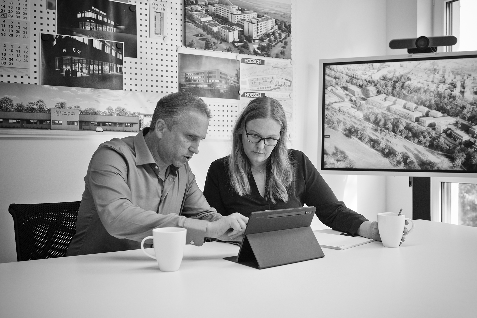 Guido Haid im Gespräch mit einer Mitarbeiterin sitzend an einem Tisch vor einem Tablet, im Hintergrund eine Pinwand mit Architekturfotos sowie ein Konferenzbildschirm mit einem Luftbild einer Wohnanlage
