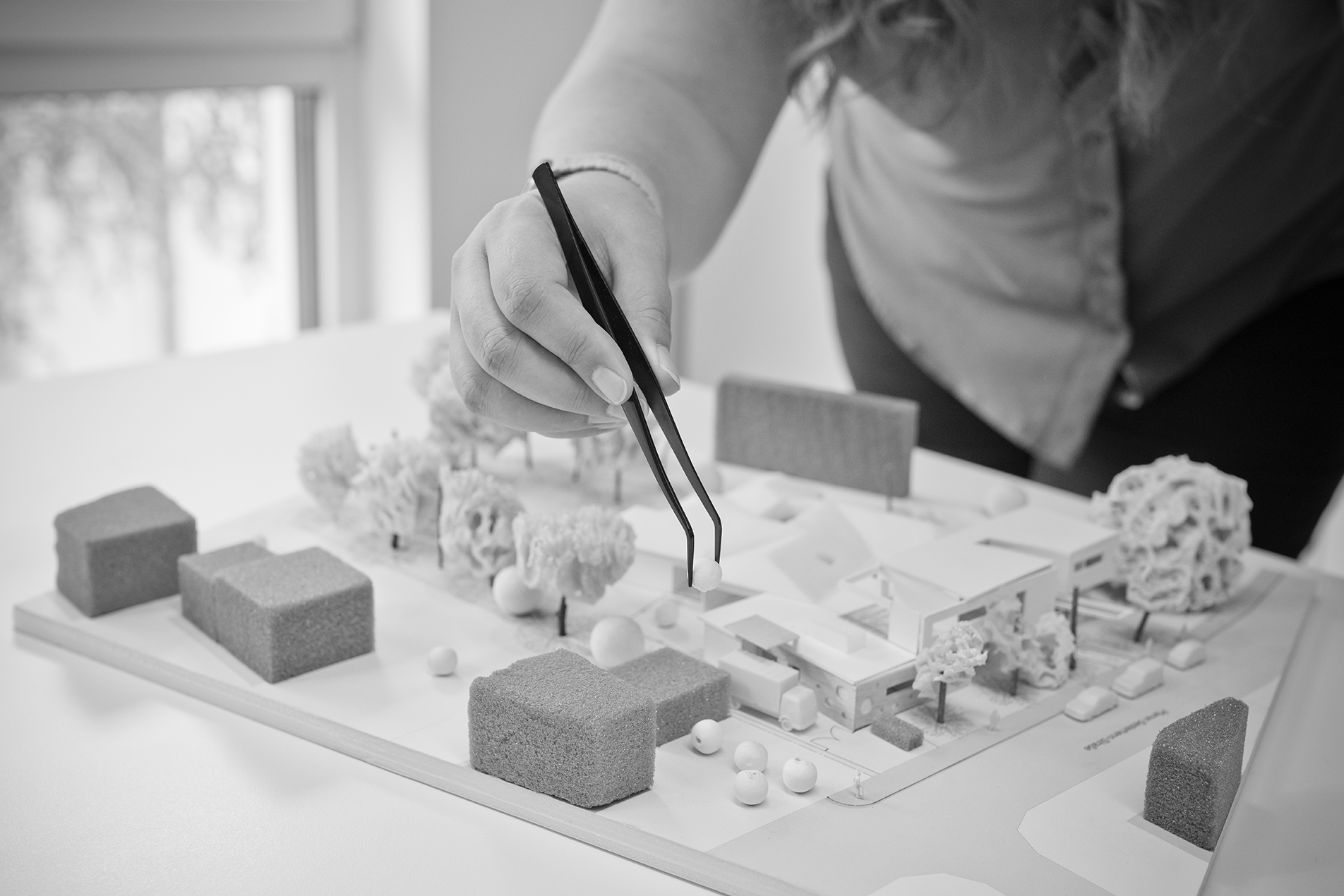 Detailaufnahme der Hand einer Mitarbeiterin, die mittels Pinzette ein Bauteil auf einem Architekturmodell platziert.